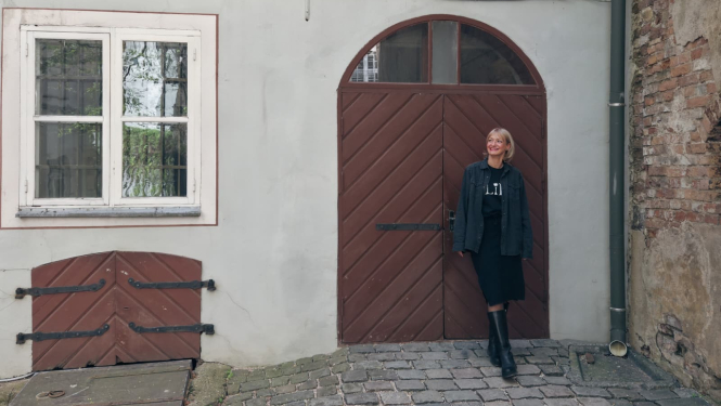Pie vēsturiskas ēkas koka durvīm stāv gaišmataina sieviete tumšās drēbēs. Pa kreisi redzams neliels četru rūšu logs un pagraba lūka, pa labi - mūra siena. Zemi klāj bruģis.