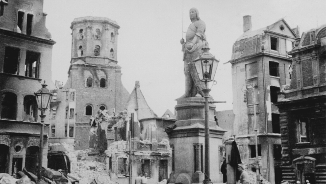 Melnbalta fotogrāfija, kurā redzams Otrā pasaules kara postījumu skats Rīgas Rātslaukumā. Attēlā redzama sagrauta pilsētas ainava ar gruvešiem un bojātām ēkām. Centrā izceļas Svētā Pētera baznīcas sagrautais tornis ar atklātām konstrukcijām, bet priekšplānā, starp iznīcinātām ēku fasādēm. Necietusi redzama Rolanda statuja. Attēls uzskatāmi demonstrē plašus pilsētas iznīcināšanas apjomus.