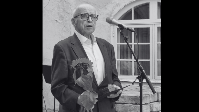 Melnbaltā attēlā ar melnām malām redzams kungs uzvalkā ar brillēm. Fonā ir ēkas logs, priekšā mikrofons. Kungs saņēmis saulespuķi un rokā ir maza kastīte, kurā ir apbalvojums. Attēls veikts svinīgā ceremonijā. Attēla melnbaltais noformējums saistīts ar to, ka attēls ilustrē līdzjūtības ierakstu.