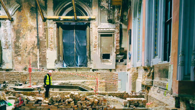 Vēsturiska ēka, kurā notiek vērienīgi būvdarbi. Tālumā redzams logs ar smailu arku, uz grīdas būvgruži.