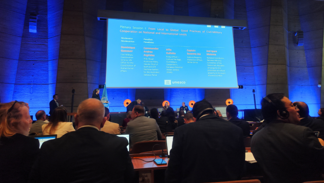 Plašā iekštelpu skatā redzama auditorija, kas sēž zālē un skatās uz lielu projekcijas ekrānu. Ekrānā ir balts teksts uz zila fona, kurā angliski norādīta plenārsesija: "No vietējā uz globālo: civilmilitārās sadarbības labā prakse nacionālā un starptautiskā līmenī". Zem virsraksta ir uzskaitīti runātāji un viņu tituli vai organizācijas. Apakšā labajā pusē ir redzams UNESCO logotips. Auditorija vērsta pret ekrānu, var redzēt daudzu cilvēku galvas un plecus. Tie sēž pie galdiem un klausās prezentāciju.  