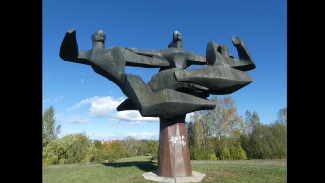 Attēlā redzams liels, tumša metāla abstrakts piemineklis, kas attēlo vairākas stilizētas cilvēku figūras, kuras savijušās kopīgā kompozīcijā un paceltas gaisā. Piemineklis novietots uz masīva pjedestāla brīvā dabā, apkārt aug koki un redzama pļava. Apakšējā daļā uz pamatnes redzams balts grafiti uzraksts. Fonā zilas debesis ar baltu mākoņu joslu.