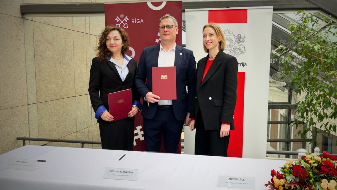 Priekšplānā atrodas galds, tas klāts ar baltu galdautu. Labajā apakšējā stūrī atrodas ziedu kārtojums, bet uz galda izkārtotas vārdu zīmītes. No labās puses - Ināra Bula, Viesturs Kleinbergs, Agnese Lāce. Visas trīs minētās personas stāv aiz galda un pozē bildei. Visi saģērbušies melnās žaketēs. Divi no cilvēkiem tur sarkanas mapes ar zeltītu Rīgas ģerboni. Aizmugurē redzami Rīgas pašvaldības un Kultūras ministrijas baneri un stikla siena. 