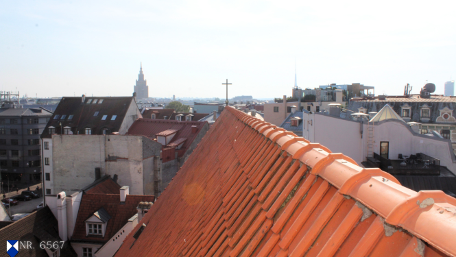 Attēlā redzams Rīgas Svētā Jāņa baznīcas jumta kores fragments tuvplānā. Jumts klāts ar sarkaniem dakstiņiem, kas veido izteiksmīgu tekstūru ar paralēlām līnijām. Jumta kores līnija stiepjas no attēla apakšējās labās puses uz augšējo kreiso pusi. Kores galā, attēla centrālajā daļā, pret debesīm paceļas tumšs metāla krusts. Fonā paveras plašs Vecrīgas panorāmas skats ar dažādu ēku jumtiem, kas ir galvenokārt sarkanos un pelēkos toņos. Fonā izceļas augsta, gaiša ēka ar smailu torni – Rīgas Zinātņu akadēmija. 