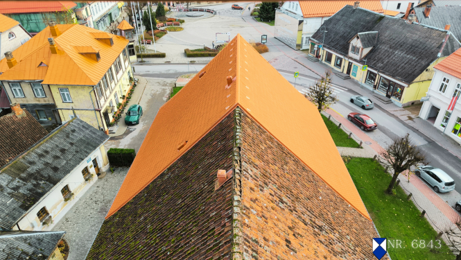 Skats no augšas uz Tukuma luterāņu baznīcas jumtu, kura viena puse nesen atjaunota ar spilgti oranžiem dakstiņiem, bet otra puse – vecāka, tumšāka, ar sūnām apaugusi. Baznīca atrodas pilsētas centrā, apkārt redzamas vēsturiskas ēkas ar dažādu veidu jumtiem, iela ar dažiem automobiļiem, gājēju ietves un koki. Attēla labajā apakšstūrī redzams kultūras pieminekļa simbols zili baltā krāsā un objekta numurs – 6843.