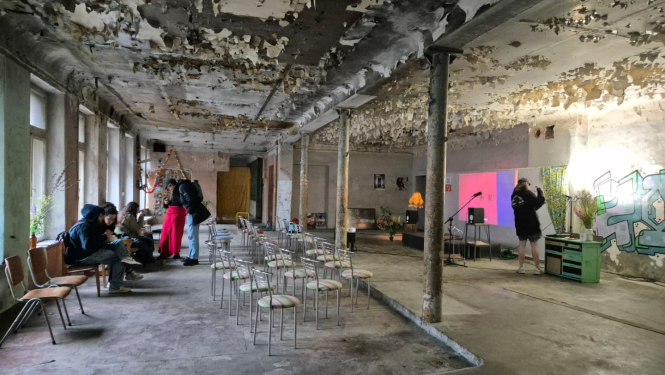 Attēlā redzami studenti. Tie atrodas plašā, neapdzīvotā vēsturiskā telpā. Tai no sienām un griestiem lobās krāsa, izskatās, ka tā tiek izmantota pagaidu pasākumiem. Grīda ir betona. Telpas vidū ir 15-20 metāla krēsli, kas vērsti ar muguru pret logiem. Telpas dziļumā ir neliela skatuve ar 2 mikrofoniem un projektoru, kas projicē rojicē gaiši rozā-purpursarkanu attēlu uz baltas sienas. Skatuves labajā pusē ir grafiti uzraksts, un uz grīdas ir novietotas puķu vāzes. 