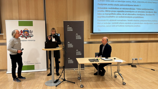 Attēlā redzama konferenču telpa koka interjerā. Telpā redzami 3 cilvēki. Pa kreisi stāv sirms kungs pelēkā džemperī un melnās biksēs. Pa vidu pie stāvgalda stāv gara sieviete ar gaišiem matiem tumšās drēbēs. Labajā pusē pie galda sēž kungs melnā uzvalkā. Aiz cilvēkiem ir Labklājības ministrijas un Arhitektu savienības baneri. Labajā sānā redzams ekrāns uz kura atspoguļojas teksts.