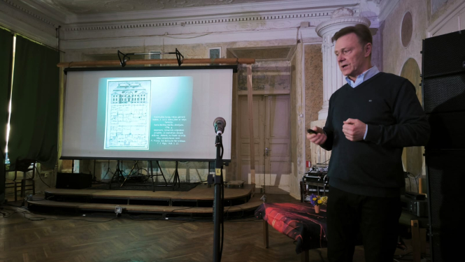 Skats ar prezentāciju. Kreisajā pusē stāv tumšās drēbēs ģērbies vīrietis ar īsiem matiem. Blakus atrodas mikrofons. Dziļumā redzama uz izvelkama ekrāna projicēta prezentācija. Viss notiek vēsturiskā telpā.