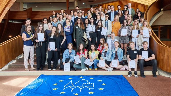 Smaidīga jauniešu un pieaugušo grupa stāv pozēdami attēlam, tiem priekšā izklāts Eiropas kultūras mantojuma dienu karogs. Karogs ir līdzīgs Eiropas savienības karogam, bet tam pa vidu ir ēku kontūras. Apkārt redzama plaša telpa ar koka interjeru.