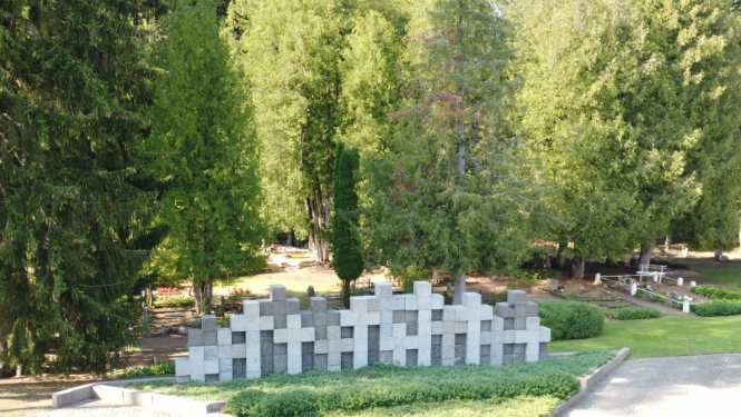 Piemiņas vieta ar kapiem, plašs lauks ar baltiem dažādu izmēru krustiem centrā. Aiz krustiem zaļo koki.