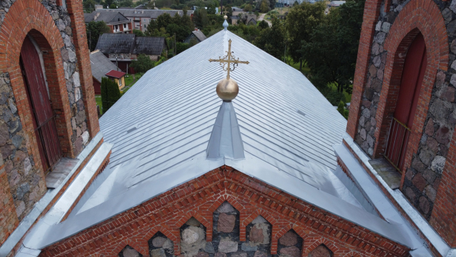 Attēlā redzams restaurētas baznīcas jumta fragments no putna lidojuma perspektīvas. Galvenajā plānā ir liels, lēzens divslīpju jumts, kas klāts ar gaiši pelēkām skārda loksnēm. Jumta korē pacelas metāla smaile ar zeltītu dekoratīvu lodi un krustu. Abās pusēs jumtam piekļaujas masīvi torņi, kas būvēti no sarkanajiem ķieģeļiem un neapstrādātiem laukakmeņiem, kuros redzamas arkveida logu ailes. Fonā pavīd nelielas ciemata mājas un koki, uzsverot objekta nozīmību vietējā ainavā.