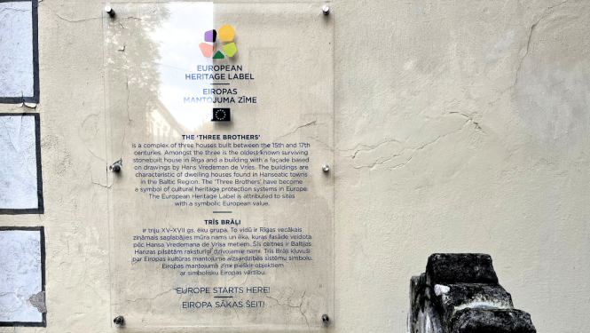 Attēlā redzama pie ēku ansambļa "Trīs brāļi" piestiprinātā Eiropas mantojuma zīme ar aprakstu. Zīme ir no stikla, lai pilnībā neaizsegtu vēsturisko ēku un būtu mazāk uztverama vidē, pati zīme ir krāsaina, bet burti - tumši zili.