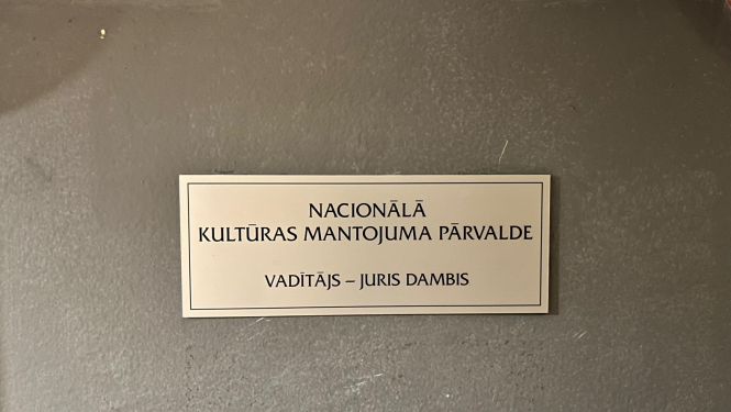 Plāksnīte pie Nacionālās kultūras mantojuma pārvaldes vadītāja durvīm, kas saka "Nacionālā kultūras mantojuma pārvalde"; vadītājs - Juris Dambis. Krāsotās koka durvis ir pelēcīgas, plāksne četrkantīga, neliela, metāla ar melniem burtiem.