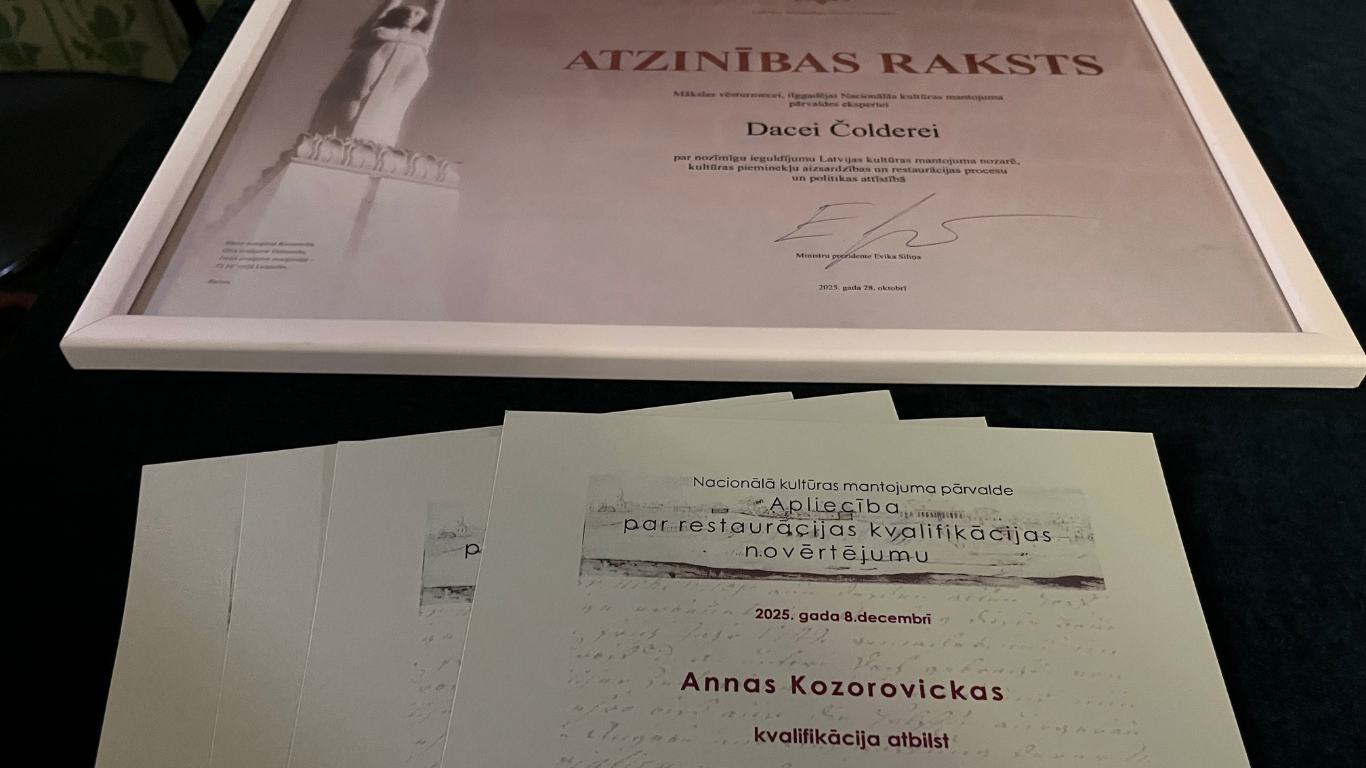 Tuvplāna foto, kurā redzams Ministru kabineta Atzinības raksts baltā rāmī un vairāki restoratora kvalifikācijas apliecinošie dokumenti zem tā.