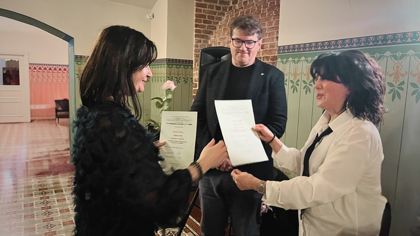 Sieviete melnā, pūkainā kleitā saņem diplomu no sievietes baltā kreklā un melnā kaklasaitē. Vīrietis melnā uzvalkā fonā tur rozā rozi.