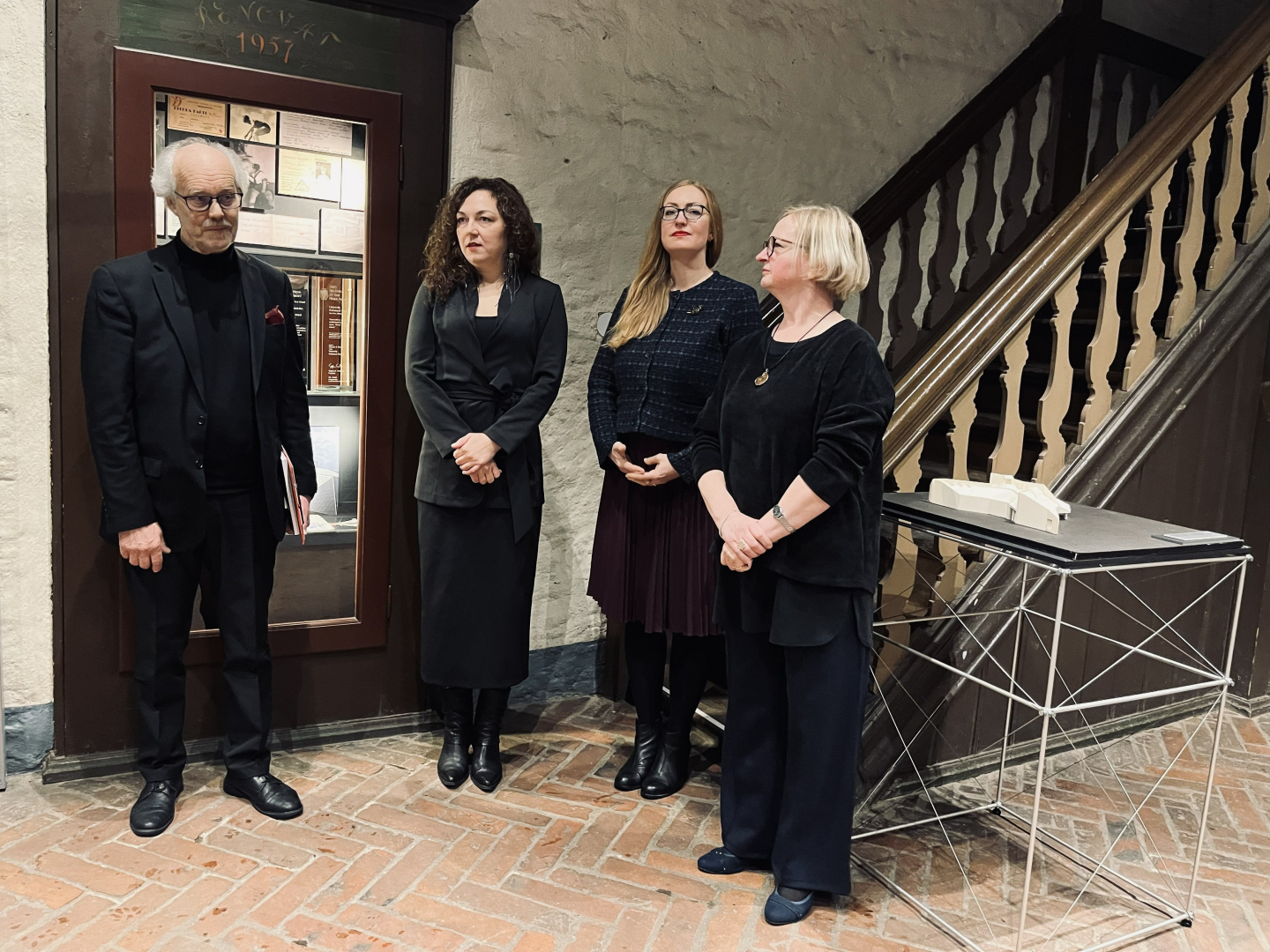 Četri pieaugušie – vīrietis sirmos matos melnā uzvalkā un trīs sievietes tumšā, lietišķā apģērbā – stāv rindā pie vēsturiskām koka kāpnēm un vitrīnas. Viņu sejas izteiksmes ir svinīgas un uzmanīgas. Vēsturiskajā telpā ir sarkanu ķieģeļu grīda un gaišas sienas. Vienai no sievietēm aiz muguras, blakus kāpnēm uz neliela galdiņa stāv makets, kas ir daļa no izstādes.