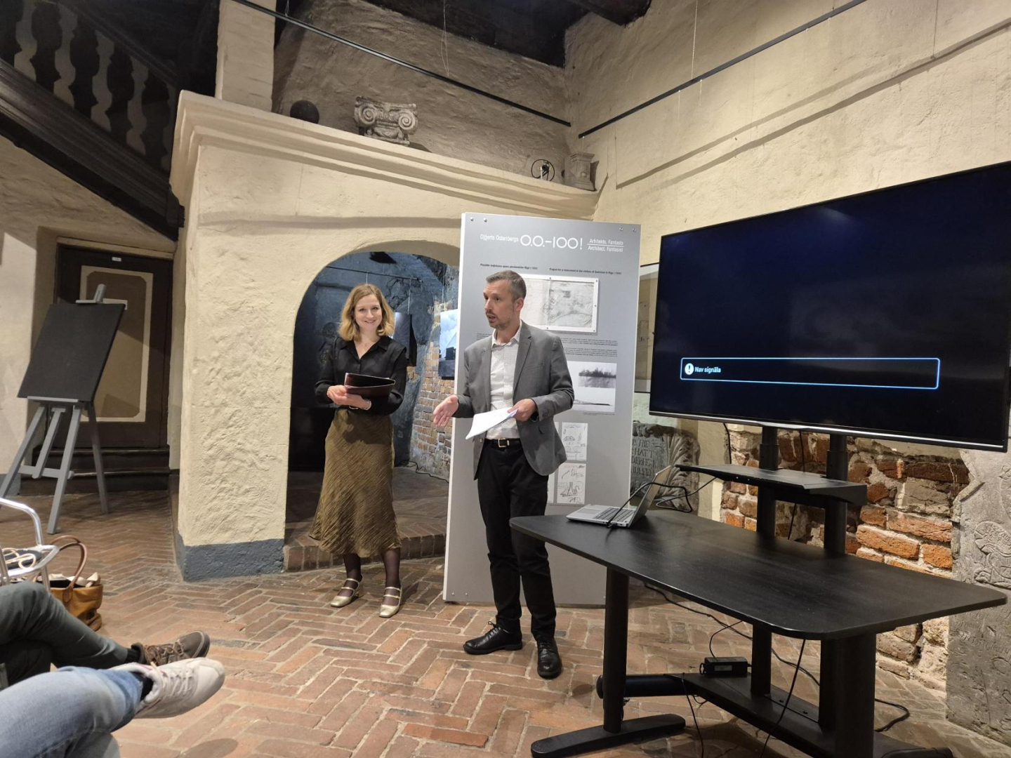 Attēlā redzama vēsturiska telpa ar sarkanu ķieģeļu grīdu, balti apmestām sienām. Vidū runā divi cilvēki - vīrietis un sieviete, pa labi redzams tumšs ekrāns, bet kreisajā pusē var manīt cilvēku kājas, kas parāda, ka telpā sēž auditorija. 