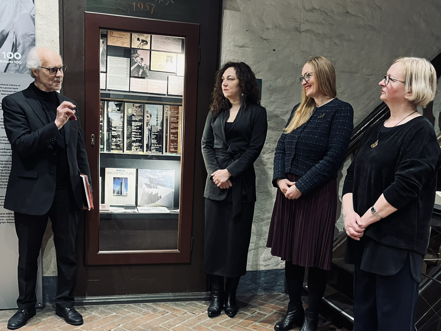 Attēla kreisajā pusē kungs sirmiem matiem, melnā žaketē ar brillēm aktīvi žestikulējot runā, vēršoties pie trim sievietēm. Sievietes stāv rindā attēla labajā pusē, ir svinīgi ģērbušās tumšās drēbēs. Visas trīs rokas tur sev priekšā sakrustotas.  Fonā vēsturiska mūra siena un vitrīna ar izstādes materiālu. Apakšā redzama sarkanu ķieģeļu grīda. Telpa ir tumši apgaismota, izceļot vitrīnu.