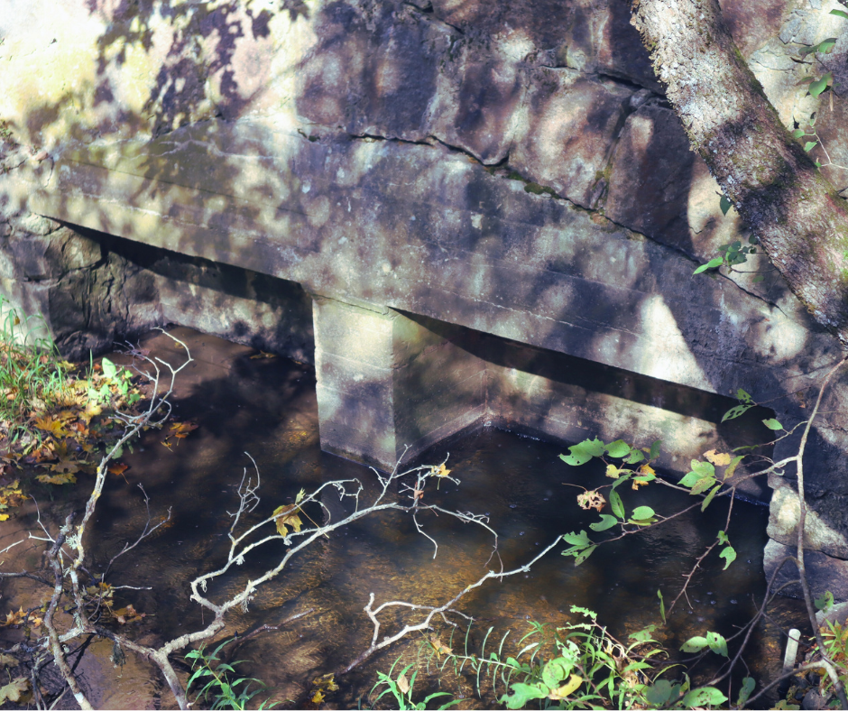 Tuvplāna attēls akmens mūra konstrukcijai, kas veido pārkares malu pār nelielu ūdensteci. Akmens siena ir tumša, ar plaisām, un tās ēnā ir maza, tīra ūdens straume. Ūdens centrā redzams gaišāks, taisnstūrveida balsts, kas atbalsta pārkari. Priekšplānā redzami zari, lapas un zaļa zāle. Visu ainu daļēji aizsedz koku ēnas un saules plankumi.