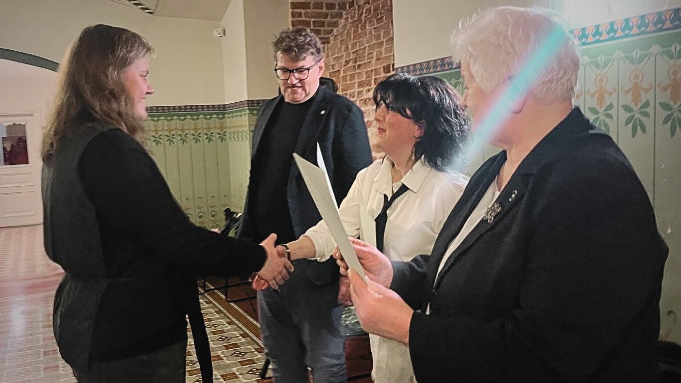 Četras personas stāv pasākuma telpā. Sievietes kreisajā pusē sniedzas rokasspiedienā ar sievieti centrā, kura tur papīrus. Vīrietis melnā uzvalkā un vecāka sieviete stāv blakus, vērojot.