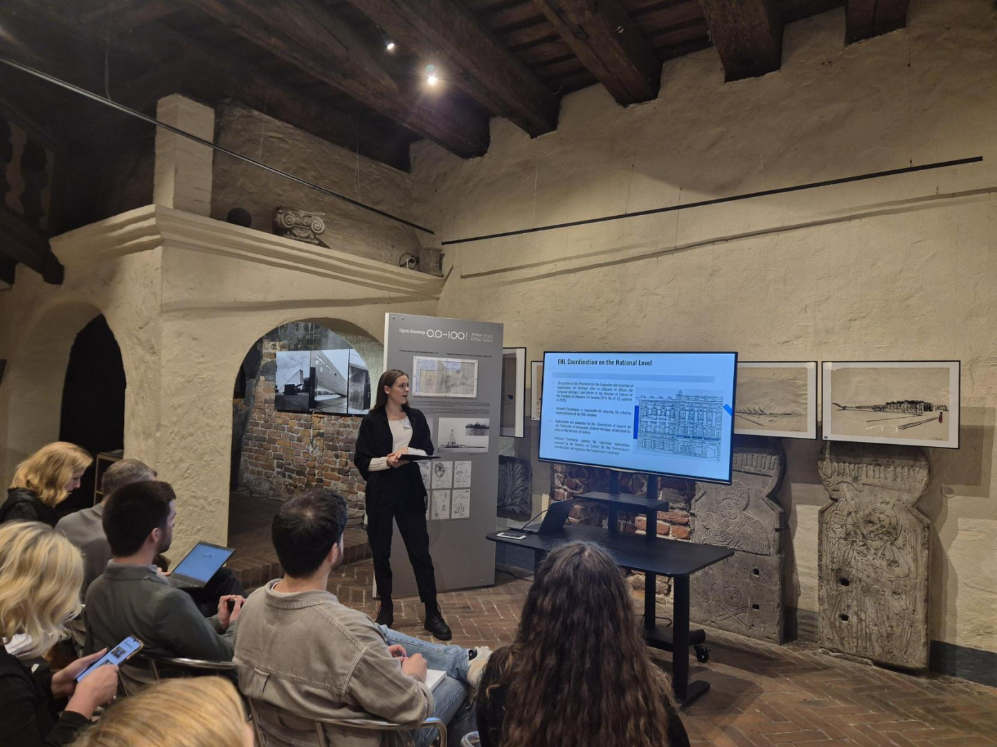 Attēlā redzama vēsturiskas ēkas telpa ar gaišām apmestām sienām, sarkanu ķieģeļu grīdu un brūnām griestu sijām. Kreisajā pusē fonā redzams apvalkdūmenis. Attēla lejasdaļā krēslos redzami cilvēki, kas klausās prezentāciju. Attēla vidū, dziļumā redzama sieviete oficiālā kostīmā, kas kaut ko stāsta un rāda uz digitālo ekrānu, kurā redzama prezentācija.