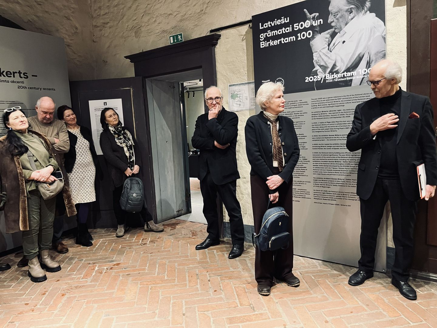 Attēla kreisajā pusē vairāki apmeklētāji stāv pie lielformāta melnbalta plakāta ar Gunāra Birkerta portretu un klausās atklāšanas uzrunas. Cilvēki ģērbušies ikdienas jakās, džemperos, dažiem plecos mugursomas. Labajā pusē redzams kungs melnā uzvalkā, sirmiem matiem, iesaistījies ekspresīvā sarunā ar kundzi, kas stāv blakus un raugās uz sarunu biedru. Kundze ģērbusies tumšās, svinīgās drēbēs, sirmiem matiem, rokās tur nelielu mugursomu. 