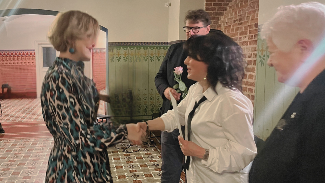 Divas sievietes rokasspiedienā pasākuma telpā ar dekoratīvām flīzēm un sienu gleznojumiem. Viena no sievietēm (kreisajā pusē) tērpta raibā kleitā, otra (centrā) baltā kreklā ar melnu kaklasaiti. Fonā stāv vīrietis ar rozā rozi.