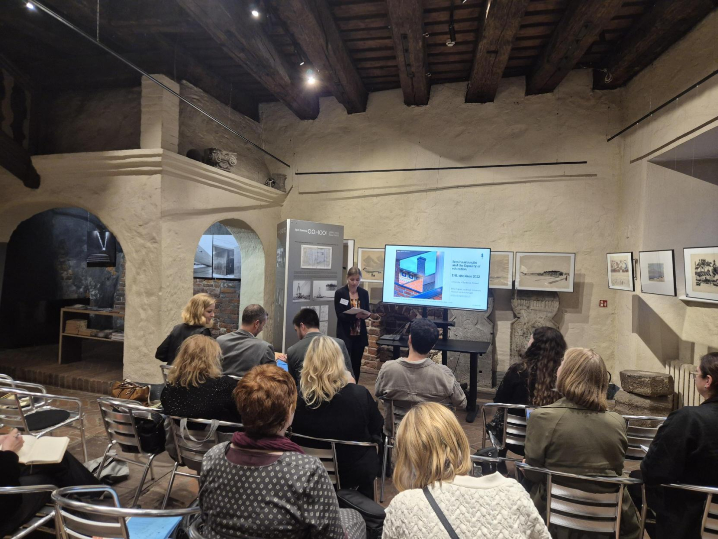 Attēls uzņemts vēsturiskā telpā ar brūniem koka griestiem un baltām, apmestām sienām. Telpā redzami krēsli, daļa no tiem ir tukši, daļā sēž cilvēki, tie fotografēti no mugurpuses. Priekšplānā ir ekrāns, uz kura tiek demonstrēta prezentācija. Prezentētāja ir sieviete tumšiem matiem, attēlā viņa ir noliekusi galvu. Telpā pie sienām karājas attāli.