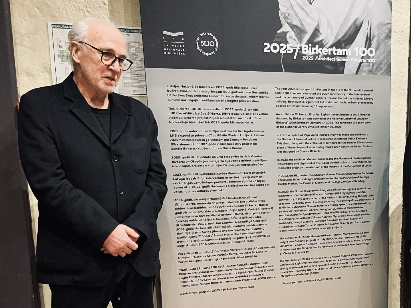 Attēla kreisajā pusē vīrietis sirmiem matiem, melnās brillēs, ģērbies tumšā uzvalkā sakrustotām rokām stāv pie informatīva stenda, kas vēstī par Gunāra Birkerta simtgadi. Apgaismojums akcentē viņa sejas vaibstus un nopietno izteiksmi