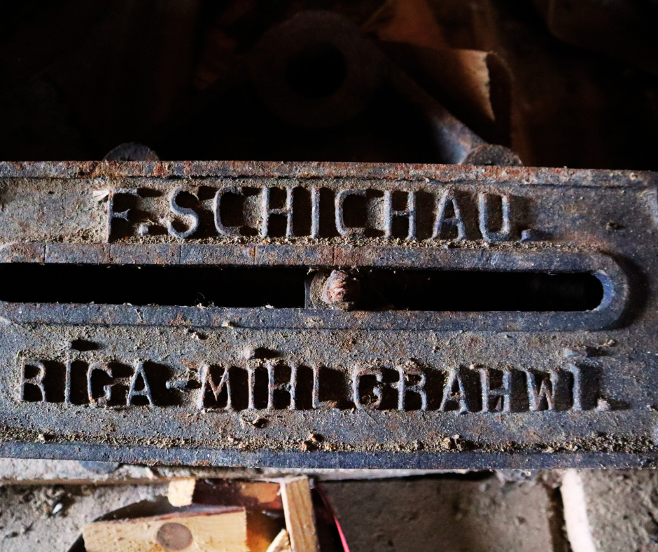 Tuvplāna attēls vecam, rūsas dzelzs mehānisma elementam. Uz detaļas ir reljefs uzraksts: augšējā rindā 'F. SCHICHAU.' un apakšējā rindā 'RIGA-MÜHLGRABEN' (Rīga-Mīlgrāvis). Starp abām uzrakstu rindām atrodas horizontāla sprauga. Detaļa ir netīra un veca, ar tumšu, raupju tekstūru.