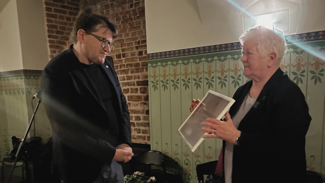 Vīrietis melnā uzvalkā stāv blakus vecākai sievietei, kura tur ierāmētu papīru (Atzinības rakstu). Vīrietis lasa sievietes rādīto papīru.Fonā redzama ķieģeļu siena un zaļi krāsotas dekoratīvas paneļflīzes.