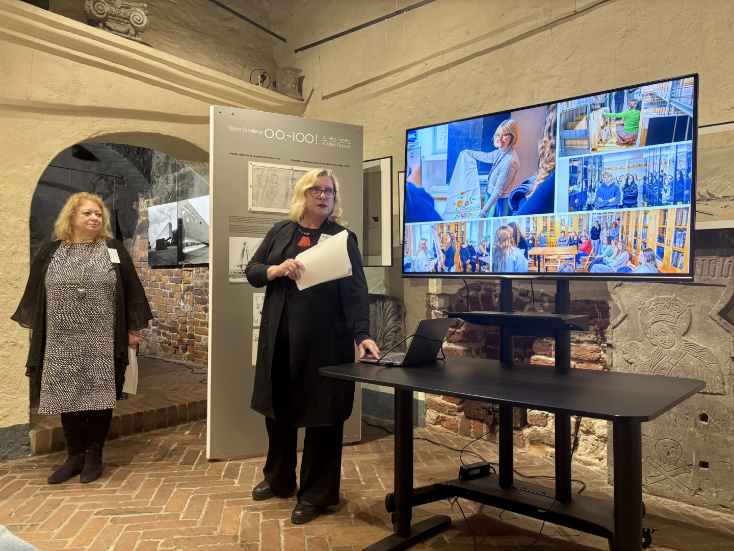 Divas sievietes stāv attēla kreisajā pusē un centrā, abas nedaudz atstatu viena no otras. Sievietēm ir gaiši mati. Abas skatās uz ekrānu, kurā redzami krāsaini attēli. Ekrāns ir fotogrāfijas labajā pusē. Pa vidu starp sievietēm fonā redzams informatīvais stends. Attēls uzņemts vēsturiskā telpā ar sarkanu ķieģeļu grīdu un gaišām, apmestām sienām