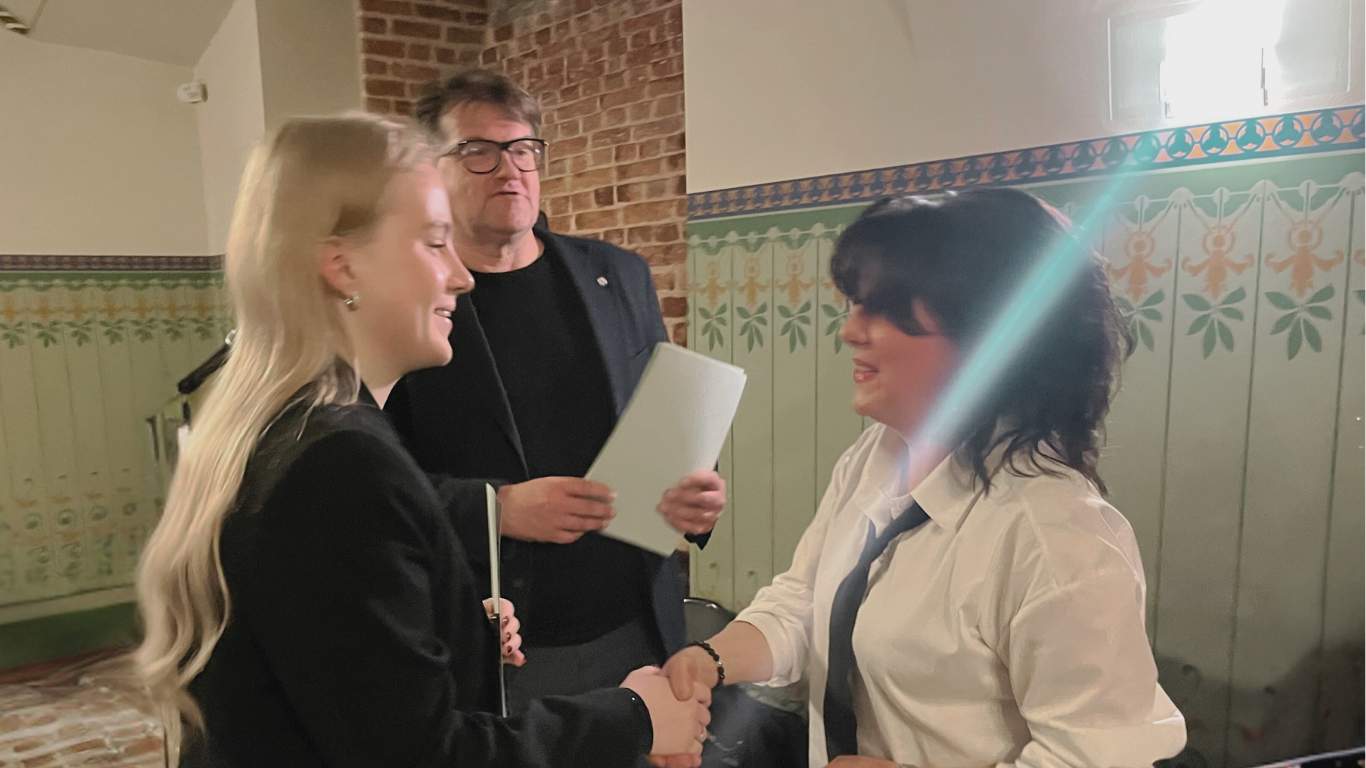 Jauna sieviete (kreisajā pusē) uzvalkā sarokojas ar sievieti (labajā pusē) baltā kreklā un kaklasaitē. Aiz viņām stāv vīrietis melnās drēbēs, turot papīrus.