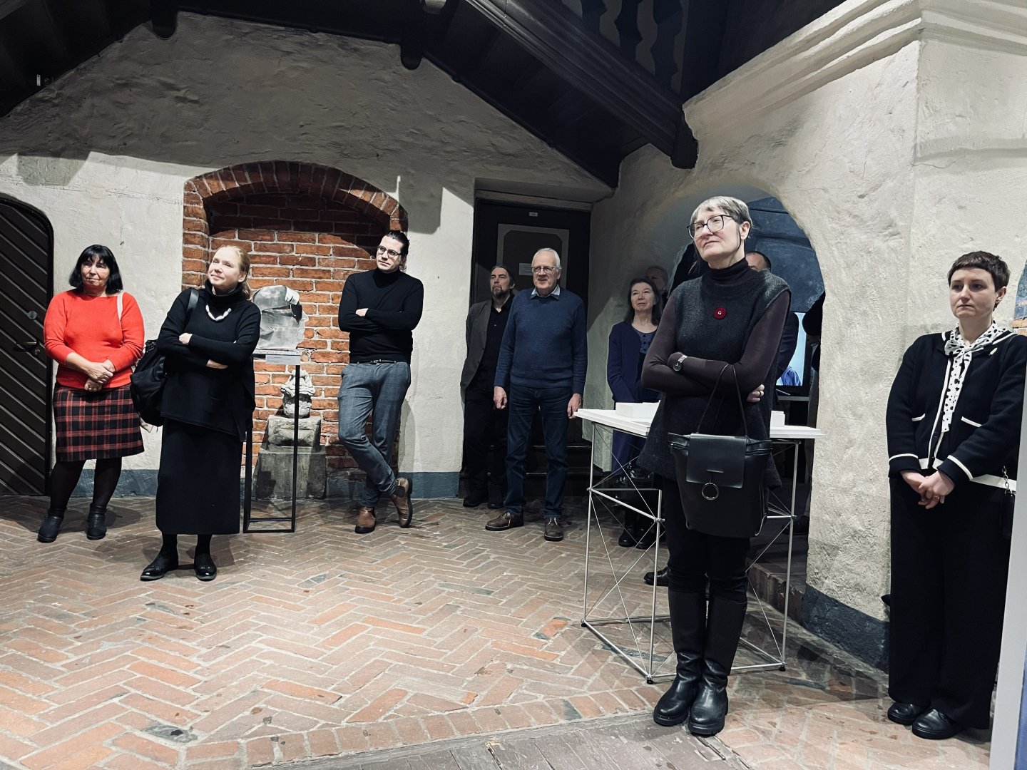 Attēlā dažādās vietās izklaidus stāv cilvēki, fonā zema mūra arka - manteļskurstenis jeb apvalkdūmenis.  Cilvēki iegrimuši domās, vērojot pasākuma norisi. Viņu vidū sievietes ar dažād matu krāsu, vīrietis tumšā džemperī. Pa vidu labi redzama sarkanu ķieģeļu grīda un starp cilvēkiem manāmi izstādes elementi.