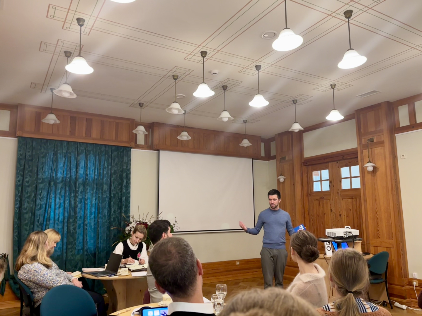 Attēlā redzama gaiša telpa. Pie gaišajiem griestiem karājas daudzas ieslēgtas lampas. Sienas ir gan gaiši krāsotas, gan ar koka paneļiem, logam priekšā aizvilkti tumši zili aizkari. Logs ir attēla kreisajā pusē un tam blakus ir izvilkts baltais ekrāns, kas paredzēts projektoram. Dziļumā centrā stāv vīrietis ar īsiem tumšiem matiem, zilā džemperī. Viņš kaut ko ekspresīvi stāsta. Attēla priekšplānā redzami cilvēki, kas sēž pie galdiem pa mazām grupām. Viņi kopīgi diskutē.