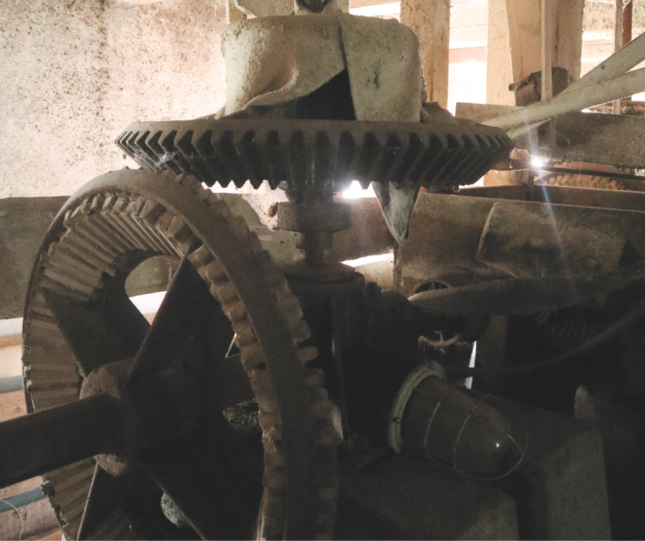 Tuvplāna attēls ar senām, putekļainām ūdensdzirnavu iekārtām. Kadra centrā atrodas zobrati. Caur  centrālā zobrata zobiem spīd gaisma no loga vai atveres, radot dramatisku efektu. Iekārtas izskatās smagas, no čuguna vai dzelzs, un ir nobirušas ar miltu putekļu slāni. 