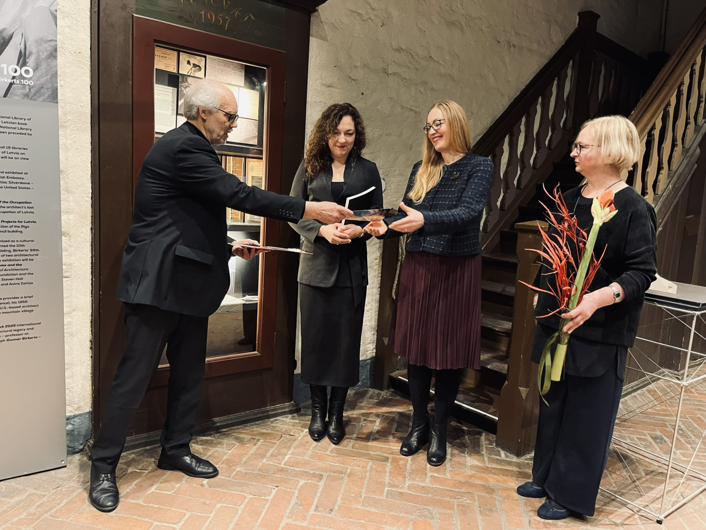 Kustības tverts mirklis: kungs sirmiem matiem, melnā uzvalkā sniedz grāmatu sievietei tumši zilā žaketē un bordo svārkos. Blakus, tuvāk attēla labajam apakšējam stūrim stāv sieviete ar sarkanu amariļļa ziedu rokās, bet aiz visiem stāv un situāciju vēro sieviete tumšiem, sprogainiem matiem. Atmosfēra ir svinīga.