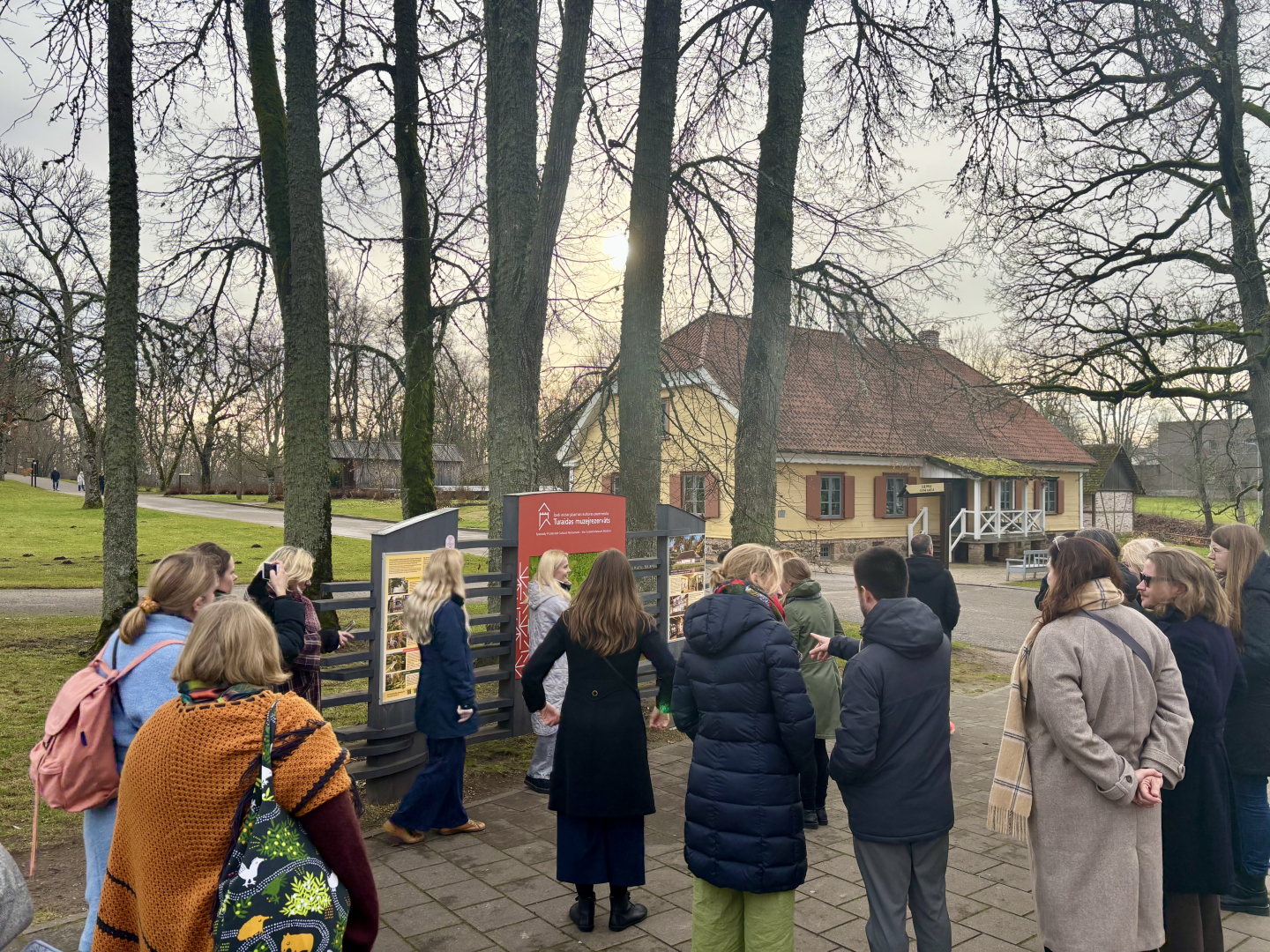 Attēls uzņemts ārā, lauku vidē, rudenī. Neliela cilvēku grupa mēteļos dodas ekskursijā. Apkārt redzami koki bez lapām un tālumā vienstāva vēsturiska māja ar divslīpju jumtu.