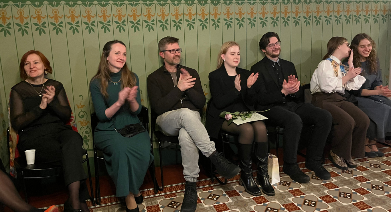 Klātesošie aplaudē sēžot sešos krēslos pasākuma zāles priekšplānā. Sienas ir dekorētas ar zaļiem zīmējumiem. Centrā redzama jauna sieviete, kura tur rokā rozā rozi.