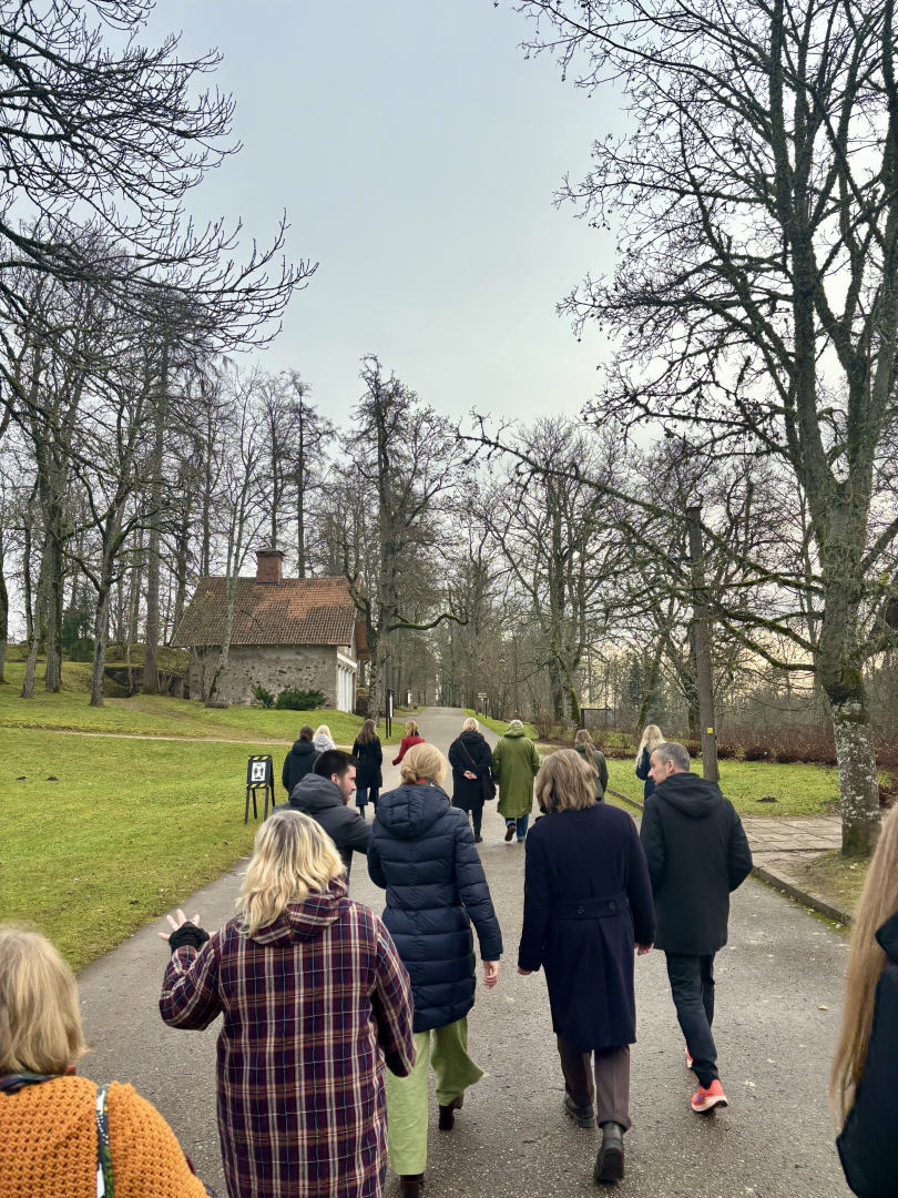 Ārtelpā uzņemta fotogrāfija, kurā redzama rudenīgi ģērbta cilvēku grupa, kas dodas ekskursijā. Apkārt redzami koki bez lapām, fonā ir zilas debesis un neliela vēsturiska ēka.