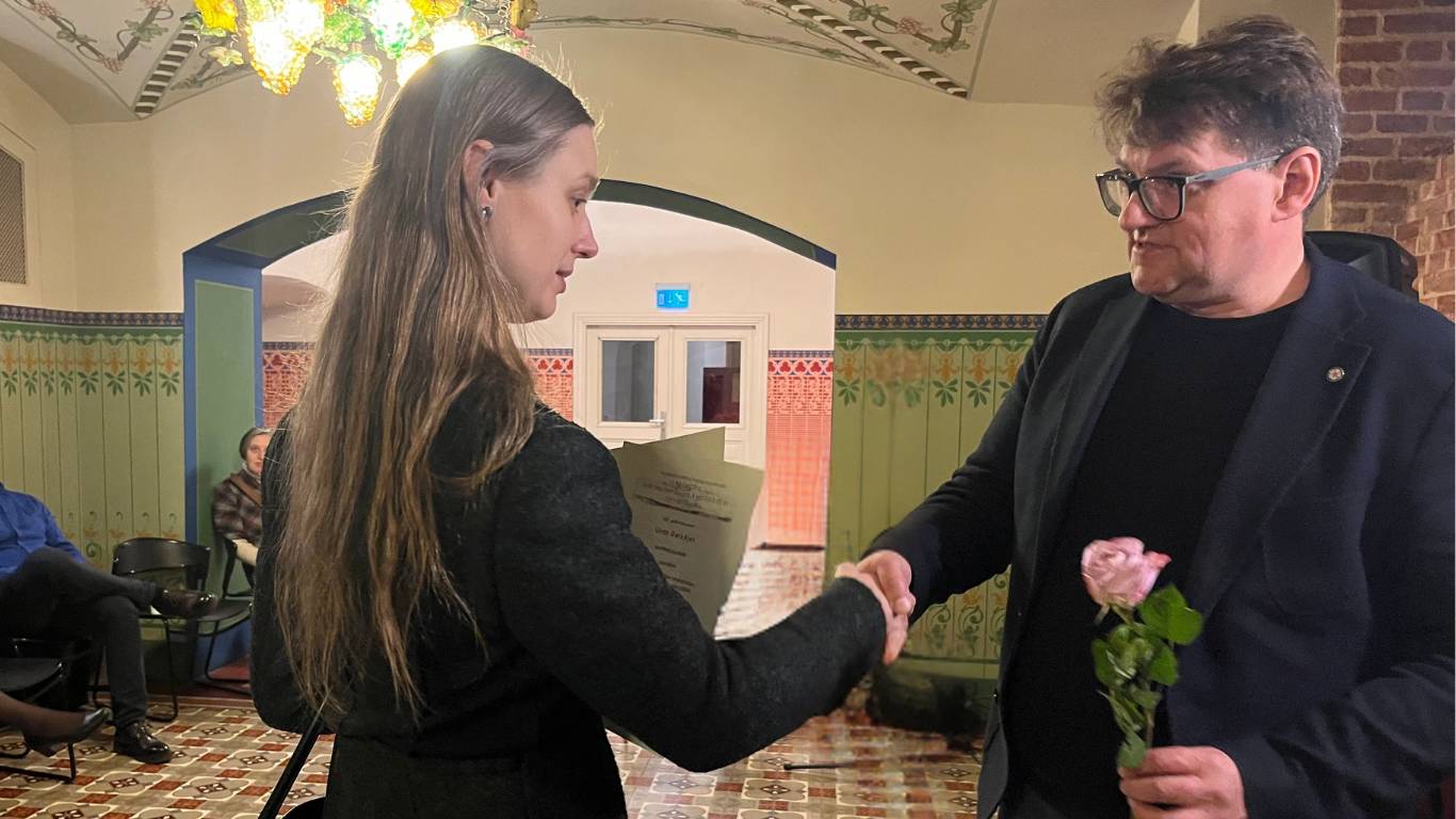 Vīrietis melnā uzvalkā un sieviete ar gariem matiem rokasspiedienā. Vīrietis tur rozā rozi. Sieviete kreisajā rokā tur diplomu.