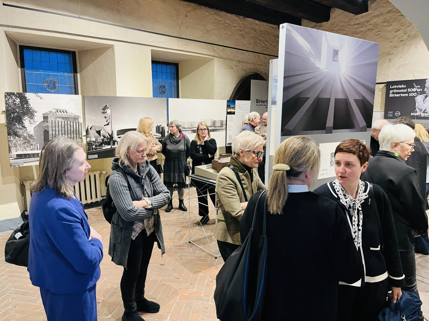Cilvēki izklaidus pa telpu tumšos un zilos apģērbos sarunājas izstāžu zālē. Fonā pie sienām izvietotas lielformāta melnbaltas arhitektūras fotogrāfijas. Telpai ir redzami vēsturiski logi ar nelielām rūtīm.
