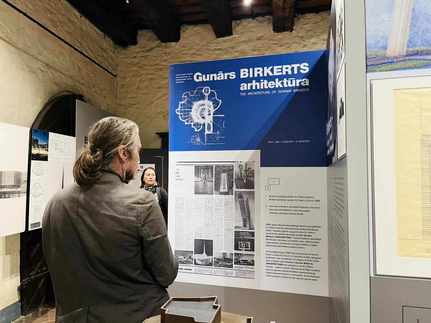 Vīrietis ar gariem, astē sasietiem pelēcīgi brūniem matiem no mugurpuses vēro lielu, zilu informatīvu stendu ar uzrakstu 'Gunārs Birkerts arhitektūra'. Uz stenda redzami ēku rasējumi un teksti.