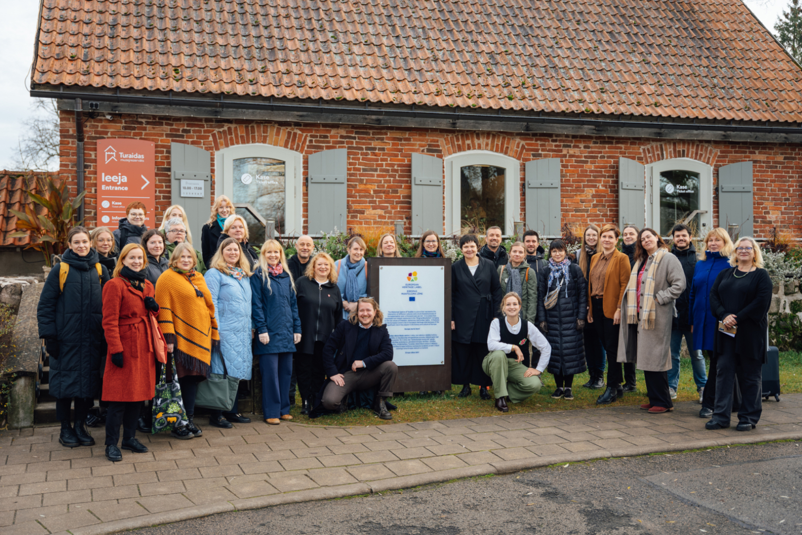 Aptveuni 30 cilvēku grupa pozē fotogrāfijai pie vēsturiskas koka ēkas ar dakstiņu jumtu. Grupa sastājusies ap lielu zīmi, kas ir Eiropas mantojuma zīme. Priekšā ir betona bruģis. Laiks ir rudenīgs.
