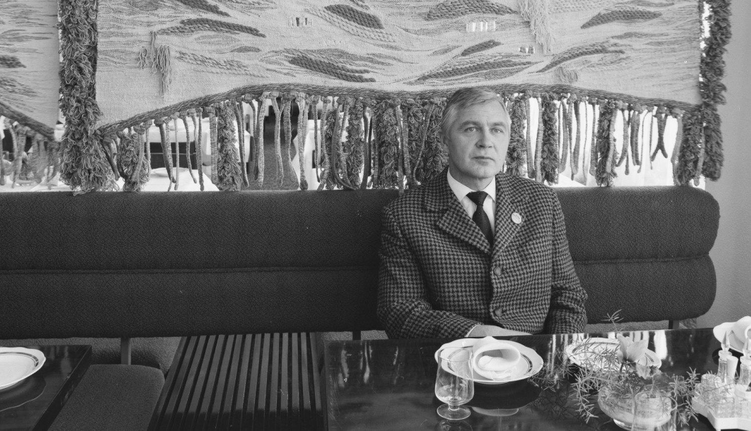 Melnbalta fotogrāfija, kurā redzams vīrietis, Oļģerts Ostenbergs, sēžot pie galda mēbelētā interjerā. Vīrietis ir redzams no krūtīm uz augšu, viņš valkā rūtainu žaketi un kaklasaiti, un skatās tieši kamerā ar nopietnu sejas izteiksmi. Aiz viņa muguras ir garš, tumšas krāsas dīvāns. Fonā aiz dīvāna redzams liels, teksturēts, iespējams, aušanas tehnikā veidots sienas dekors ar abstraktiem rakstiem un bārkstīm, kas aptver visu sienu. Pie galda priekšplānā ir redzamas šķīvju, glāžu un galda piederumu daļas.