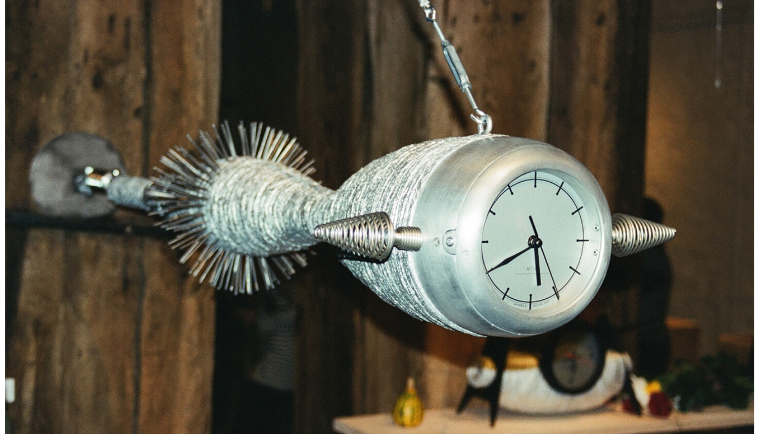 Fotoattēlā redzams dizaina pulkstenis, veidots zemūdenes vai ūdens radības formās ar sudraba krāsas korpusu un baltu ciparnīcu ar melniem rādītājiem. Aiz muguras tam redzama koka dēļu siena. Fotoattēls ir uzņemts no neliela leņķa, un gaisma ir nedaudz izkliedēta.