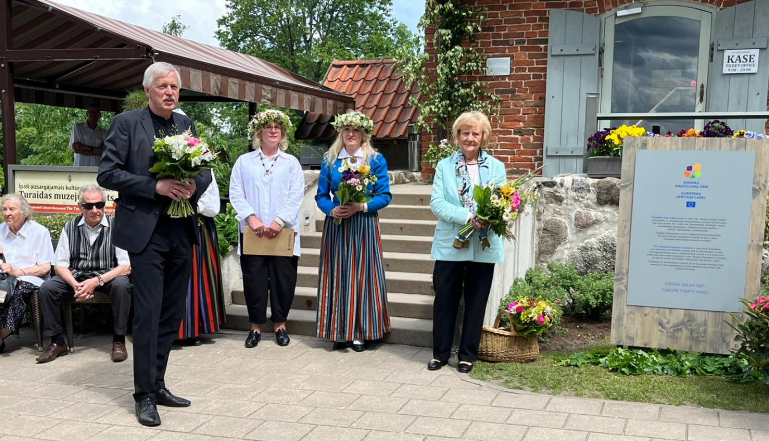 Uz betona bruģa celiņa stāv četri cilvēki - garš, sirms kungs tumšā uzvalkā ar puķu pušķi un trīs sievietes - divām galvā ir puķu vainadziņi, vidējā tērpusies latviešu tautas tērpā, sievietēm rokās ir pušķi. Cilvēki stāv blakus Eiropas mantojuma zīmes plāksnei. Fonā redzamas vēsturiskas ēkas - sarkani ķieģeļi un dakstiņi. 