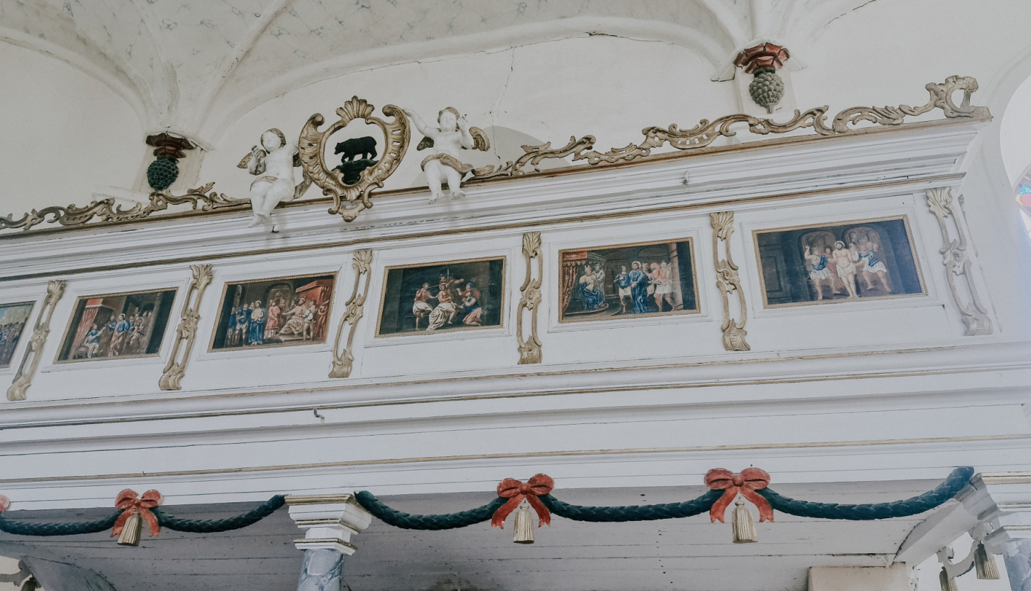 Attēls uzņemts baznīcā,  un tajā redzams balts, grezni dekorēts koka balkons. Balkona augšējā daļā ir rotājumi ar volūtām un figūrām. Centrā ir tumšs, trīsdimensionāls lācis, kas stāv uz četrām kājām. Abpus lācim sēž divas baltas, mazas eņģeļu figūriņas.  Zem augšējā rotājuma redzamas piecas gleznas ar bibliskiem sižetiem, kas ierāmētas zelta rāmjos un izvietotas rindā. Zem gleznām ir platāka koka josla, kuras apakšdaļu rotā tumši zaļa vītne ar diviem sarkaniem, dubultiem bantīšu mezgliem.  Gaismas un ēnas 