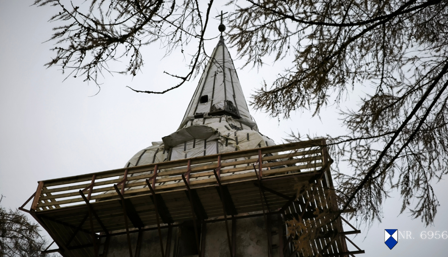 Popes luterāņu baznīcas torņa smaile remontdarbu procesā, fotografēta no apakšas. Tornim noņemts segums, to klāj balts aizsargmateriāls, bet apkārt redzamas koka sastatnes. Smailē redzams krusts un lode, fonā — lapegles zari. Attēls ieturēts tumši pelēcīgos toņos. Labajā apakšējā stūrī pievienota zilā vairoga zīme un baznīcas valsts aizsardzības numurs 6956.