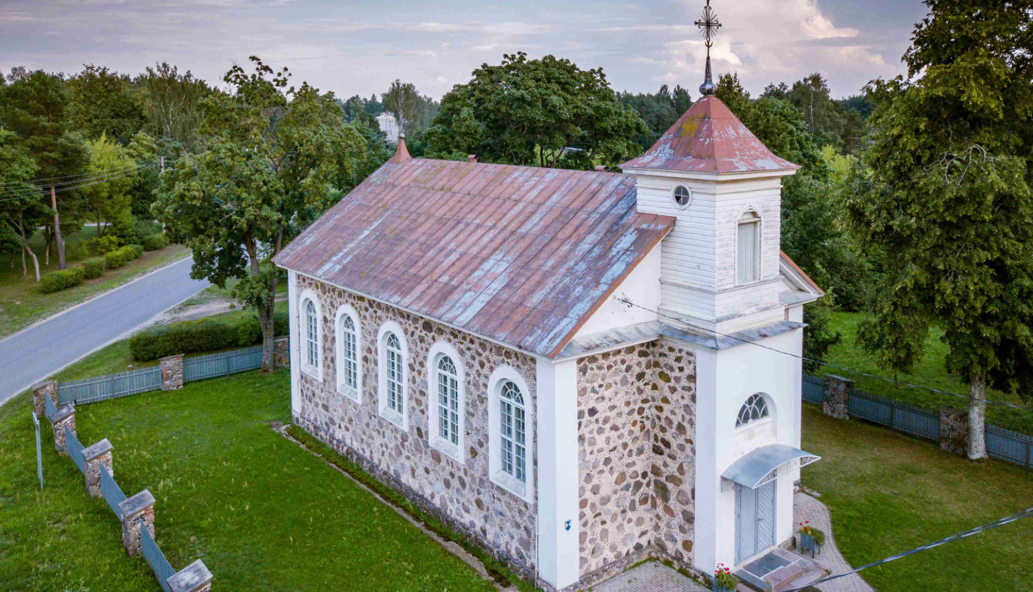 Aero foto redzama neliela, gaiša baznīca vasarīgā ainā. Mūrēta no akmens, apmestās fasādes daļas ap logiem, durvīm un nelielu tornīti, kas knapi paceļas virs divslīpju jumta kores, ir baltas. Tornīša galā - krusts. Rūsējis skārda jumts, sīkrūtiņu logi ar aploci. Baznīca ir  centrā. Labā apakšējā stūrī stiepjas celiņš, apkārt zālājs, ko ieskauj žogs, tā raksturs nav nolasāms. Kreisajā pusē asfaltēts ceļš. Debesis ir vasarīgi zilas ar mākoņiem un fonā uz visām pusēm redzami zaļi koki.
